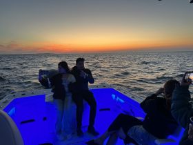 A couple captures a selfie while enjoying drinks on a boat at sunset, creating a memorable moment against a stunning ocean backdrop.