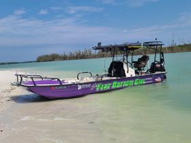 A vibrant purple flat bottom boat named 'Flat Bottom Girl' rests on a sandy shore, showcasing its sleek design against a backdrop of clear turquoise waters.
