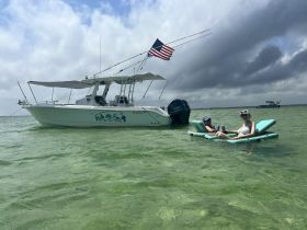 Two friends relax on floating mats in shallow water near a boat, enjoying a sunny day on the water — perfect for a fun and relaxing outing.