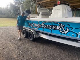 A man poses next to a vibrant charter boat named 'Team Vinson Charters,' showcasing its sleek design and branding. Perfect for fishing and adventure enthusiasts.
