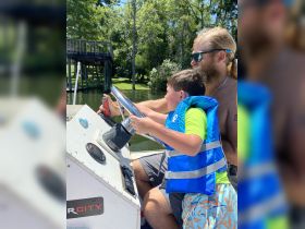 A child joyfully takes the helm of a boat with guidance from an adult, creating a memorable summer experience on the water.