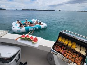A lively group enjoys a fun day on the water with a floating raft, delicious grilled food, and refreshing watermelon — perfect for a summer party at sea.