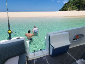 Two men enjoy a relaxing moment in the clear waters of a sandbar, with a beautiful beach backdrop — perfect for fishing and leisure activities.