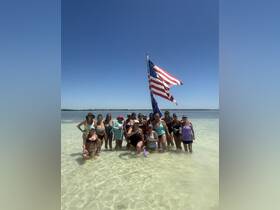 A lively group of women celebrates together in shallow waters, holding drinks and posing with an American flag — a perfect moment of fun and camaraderie at the beach.