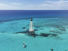 A stunning aerial view of a lighthouse surrounded by crystal-clear turquoise waters, showcasing the beauty of the ocean and nearby boats — perfect for island hopping adventures.