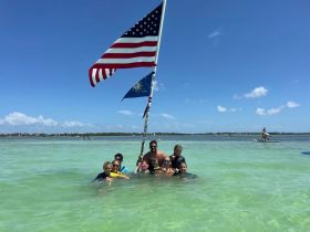 A joyful group of family and friends celebrates together in shallow waters, holding a flag at a sandbar stop — a perfect moment for fun and relaxation under the sun.