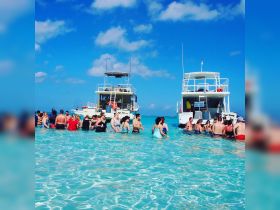 A lively group enjoys a sandbar stop, wading in crystal-clear waters with boats anchored nearby — perfect for a fun day on the water.