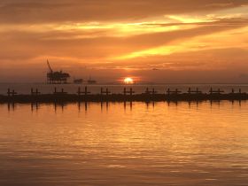 A stunning sunset over the ocean, with silhouettes of oil rigs and a pier in the foreground, creating a picturesque coastal scene.