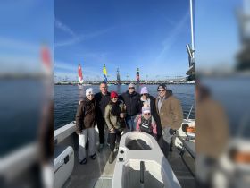A cheerful group of family and friends poses on a boat, enjoying a beautiful day on the water with colorful sails in the background — perfect for memorable outings and celebrations.