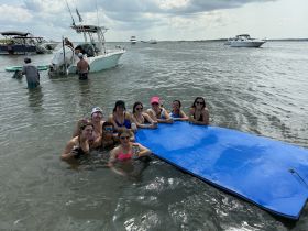 A lively group of friends enjoys a fun day in the water, relaxing on a floating mat while surrounded by boats — perfect for summer parties and memorable outings.