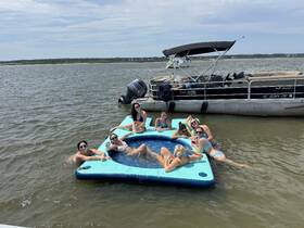 A lively group enjoys a fun day on the water, relaxing on a floating mat while sipping drinks near a pontoon boat — perfect for summer gatherings and celebrations.