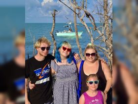 A cheerful group of friends poses for a photo by the beach, with a boat visible in the background, capturing the essence of a fun day on the water.