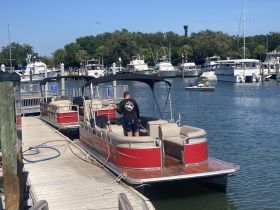 A person cleans the interior of a pontoon boat at the dock, showcasing the comfortable seating area — perfect for family outings or group gatherings on the water.