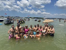 A lively group enjoys a fun day at the sandbar, swimming and celebrating with drinks, surrounded by boats and jet skis — the perfect summer getaway.