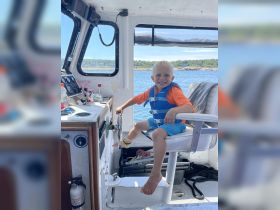 A cheerful child in a life jacket sits at the helm of a boat, ready to steer. This cozy cabin setting showcases the excitement of young adventurers on the water.