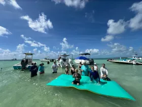 A lively group enjoys a sandbar stop, playing on a floating mat in shallow waters — perfect for family fun and summer adventures.