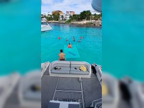 A lively group enjoys swimming and playing on a floating mat in the clear turquoise waters, showcasing the perfect day for water activities and fun in the sun.