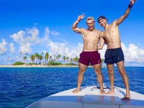 Two friends pose joyfully on the bow of a boat, with a picturesque island backdrop — a perfect moment capturing the spirit of adventure and fun on the water.