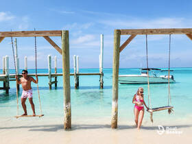 A couple enjoys a sunny day at the beach, swinging on wooden swings with a boat anchored in the background — a perfect tropical getaway scene.