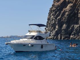 A scenic view of a boat anchored near a rocky coastline, with a group enjoying the water on a floating mat — perfect for summer adventures and beach outings.
