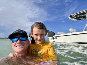 A joyful moment captured as a man and a girl swim together in clear waters, with a boat visible in the background — perfect for family adventures on the water.