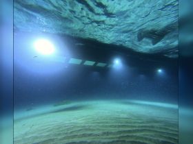 An underwater shot featuring the underside of a boat with lights illuminating the sandy bottom — the scene captures the serene underwater environment.