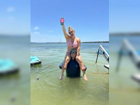 A joyful moment captured as a woman playfully poses on a man's shoulders in shallow water, celebrating a fun day at the beach with drinks in hand.