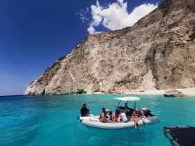A lively group enjoys a day on the water, relaxing on a boat near a stunning beach and rocky coastline, perfect for summer adventures.