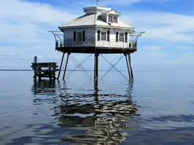 A unique structure stands on stilts over calm waters, reflecting the serene sky — an intriguing sight for those exploring coastal architecture.