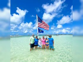 A joyful family poses together in shallow water, celebrating their day at the beach with an American flag in the background — a perfect snapshot of summer fun and togetherness.
