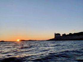 A stunning sunset over the coastline, with a lighthouse silhouetted against the vibrant sky — a perfect moment for capturing the beauty of nature while on a boating adventure.