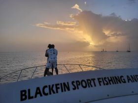 A couple enjoys a romantic moment on the bow of a boat, watching a stunning sunset over the water — an ideal setting for unforgettable fishing trips or sunset cruises.