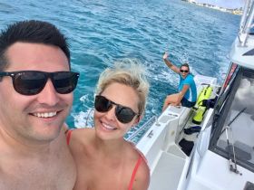 A cheerful couple takes a selfie on a boat, with a friend waving in the background — capturing the joy of a fun day on the water.
