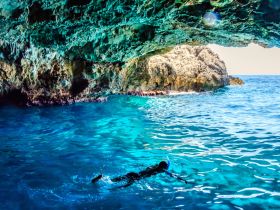 A snorkeler explores the vibrant underwater world beneath a rocky cave, showcasing the stunning turquoise waters and natural beauty of the destination.