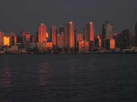 A stunning sunset casts a warm glow over the skyline, reflecting beautifully on the water — a picturesque view of the city from the harbor.