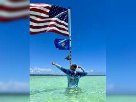 A man stands in shallow water, proudly displaying the American flag while pointing towards the horizon — a vibrant moment celebrating freedom and adventure on the water.