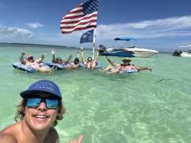 A fun group enjoys a sandbar stop, floating in the clear waters while waving and celebrating — a perfect day for relaxation and camaraderie on the water.