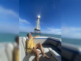 A woman relaxes on a boat while pointing towards a striking lighthouse in the distance, capturing a perfect moment of leisure on the water.