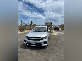 A view of a silver car parked near a gate in a desert landscape, with cacti and a cloudy sky in the background.