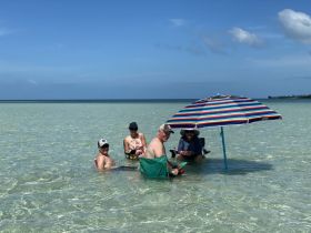 A cheerful group enjoys a sunny day at the beach, relaxing under a colorful umbrella in shallow waters — perfect for a fun day out with friends.