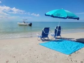 A serene view of a speedboat anchored just off the beach, with beach chairs and an umbrella set up for relaxation under the clear blue sky.