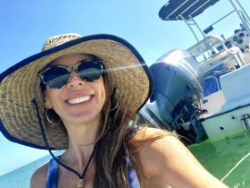 A joyful woman takes a selfie on a sunny day with a boat in the background, capturing the essence of a fun day on the water.