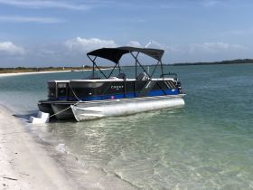 A pontoon boat named Crest is docked on a sandy beach, surrounded by clear waters, perfect for a relaxing day on the water.