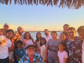A joyful group of friends and family gather for a sunset photo by the water, capturing a memorable celebration with smiles and laughter.