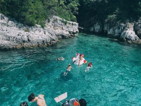 A lively group enjoys a refreshing swim in crystal-clear waters, featuring fun inflatables and stunning rocky scenery — perfect for summer adventures.