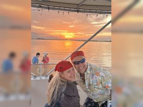 A cheerful couple dressed in pirate-themed attire smiles for the camera against a stunning sunset backdrop, capturing a memorable moment on their boating adventure.