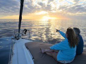 A couple enjoys a serene sunset while sailing, pointing out the beautiful horizon — a perfect moment for romantic getaways on the water.