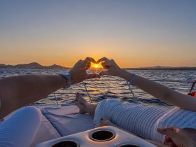 A couple forms a heart shape with their hands against a stunning sunset backdrop while relaxing on a yacht — an ideal moment for romantic getaways or special celebrations.