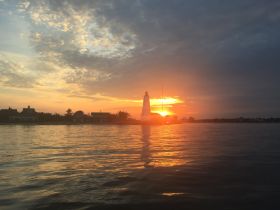 A stunning sunset casts a warm glow over a lighthouse and coastline, creating a picturesque scene perfect for evening boat trips.
