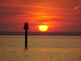 A stunning sunset casts vibrant colors over the water, with a navigational marker silhouetted against the glowing horizon — a perfect scene for evening boat trips.